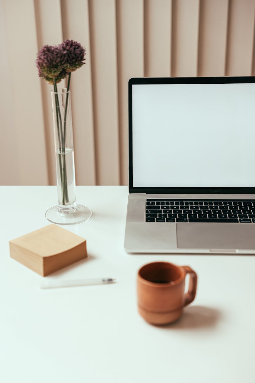 A photo of a desk that has a glass vase with a single flower in it, an open laptop, a brown mug and a stack of sticky notes. 
Photo by Cup of Couple on Pexels.com